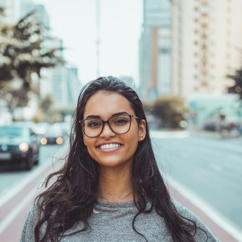 A young woman with glasses smiling on a city street, embracing urban lifestyle.
