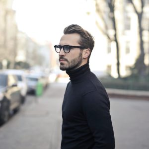 A fashionable man in a black top and glasses poses in an urban street with parked cars.
